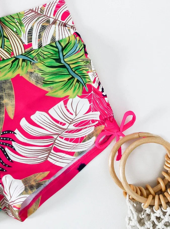 A vibrant pink and green floral print two-piece high-waist bikini set with a woven straw bag.