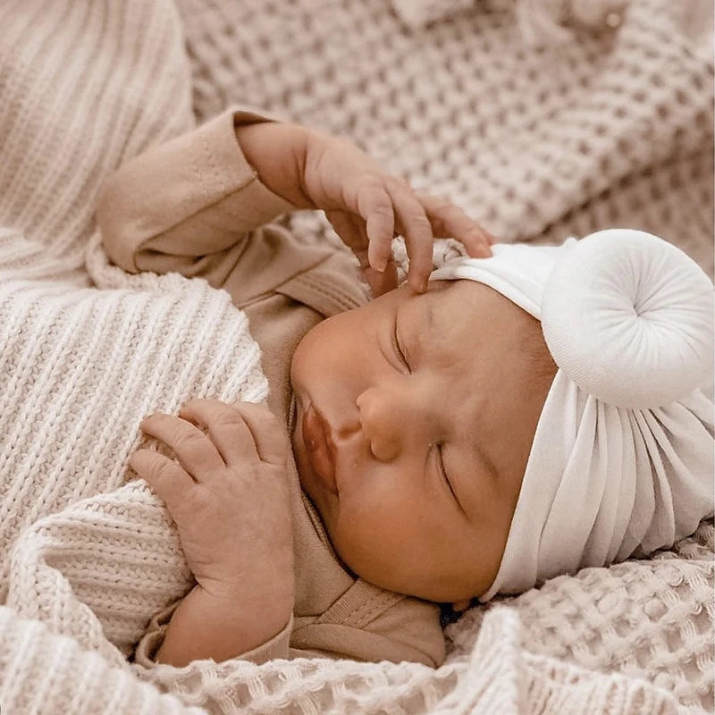 Neugeborenes Baby schläft friedlich in einem hellbeigen Baumwollturban und einer kuscheligen Strickdecke.