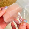 Une main tient une cacheoire de sein en silicone transparent avec un centre texturé, emballée dans une pochette en plastique 
