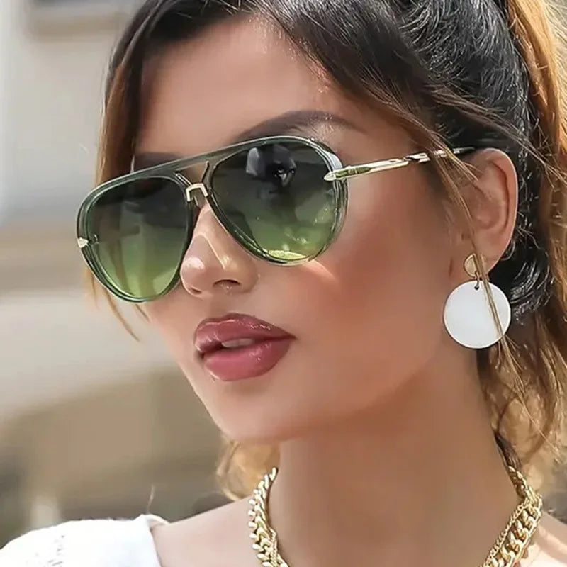 A woman wearing olive green aviator sunglasses with gold accents and a gold chain necklace.