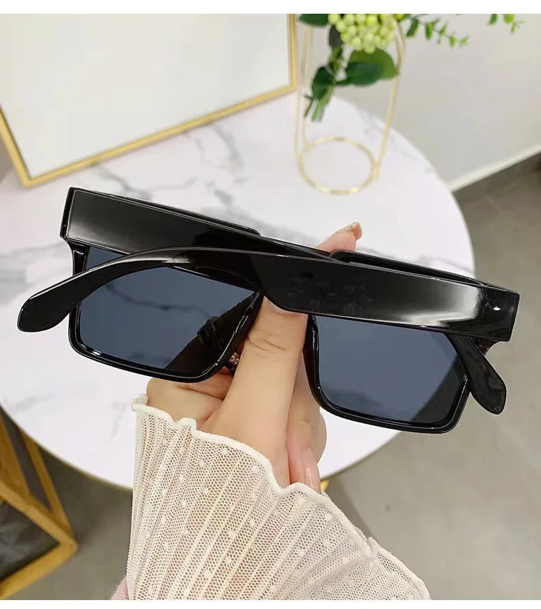 A hand holds a pair of black oversized square sunglasses against a white background.
