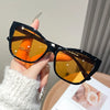 A hand holds a pair of black vintage square cat eye sunglasses with amber lenses against a light background.