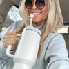 A blonde woman holds a white tumbler with a silicone straw cover and a flower on it.