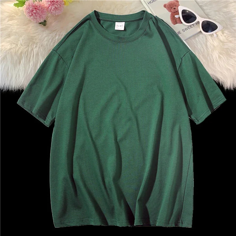 Dunkelgrifes, oversized Baumwoll-T-Shirt einer Frau auf weißem Plüsch neben Sonnenbrille und Teddybär.