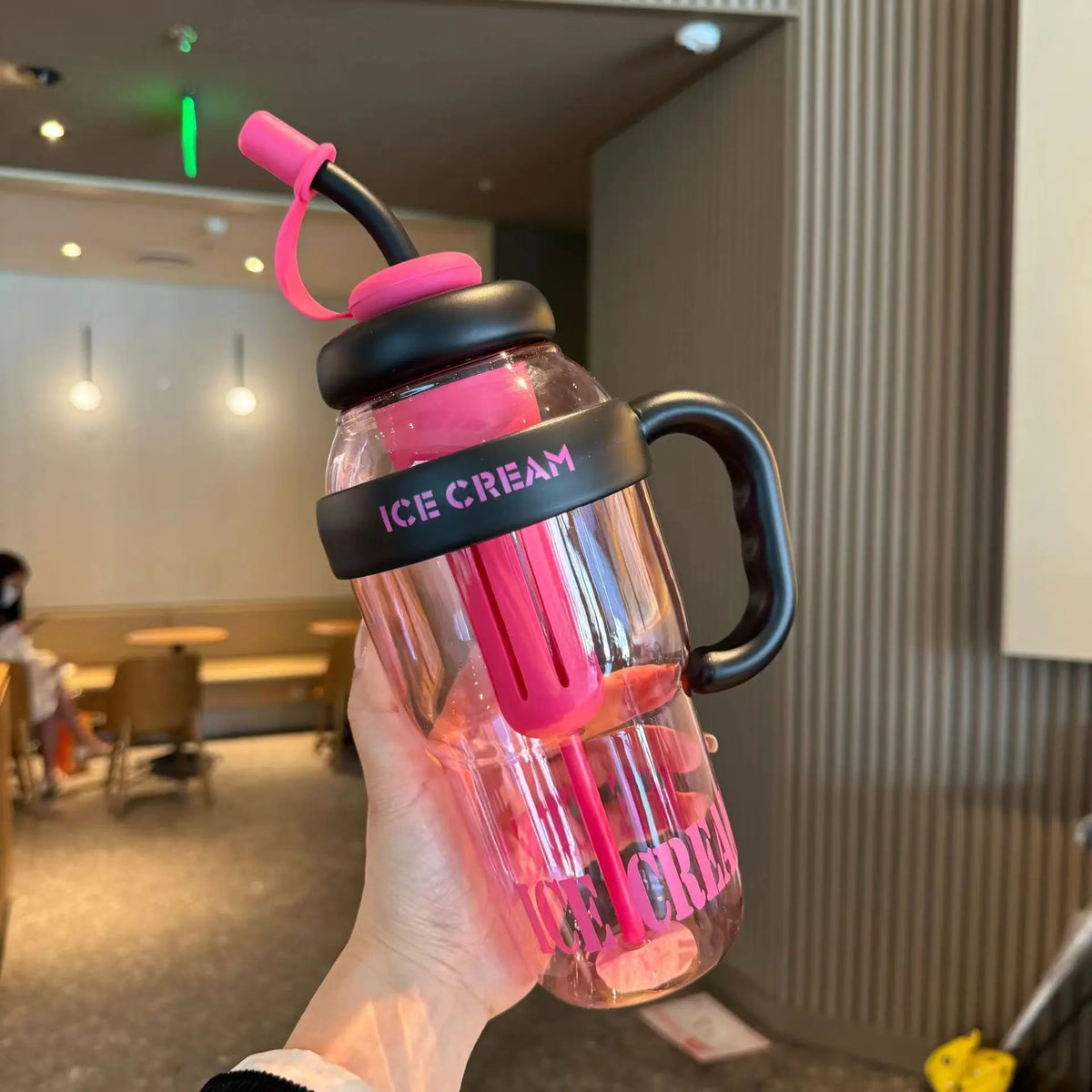 A hand holds a pink sports water bottle with a black lid and pink straw against a neutral background.
