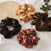 A collection of satin scrunchies in shades of gold, brown, and black are displayed on a woven mat.