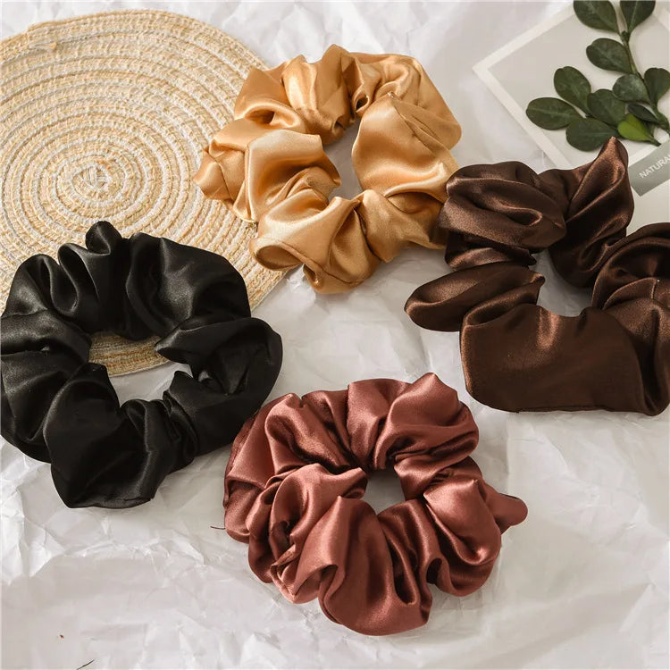 A collection of satin scrunchies in shades of gold, brown, and black are displayed on a woven mat.