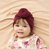 Lächelndes Baby mit maroonem Baumwollturban vor beigeem Hintergrund.