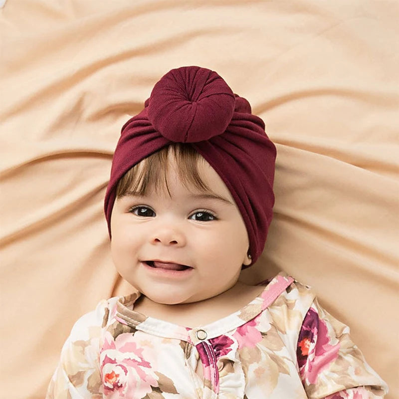 Lächelndes Baby mit maroonem Baumwollturban vor beigeem Hintergrund.