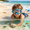 A young child wearing blue swimming goggles is looking at colorful fish in clear ocean water on a sandy beach.