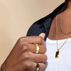 A hand wearing a gold ring and a gold necklace is visible against a dark shirt and white top.