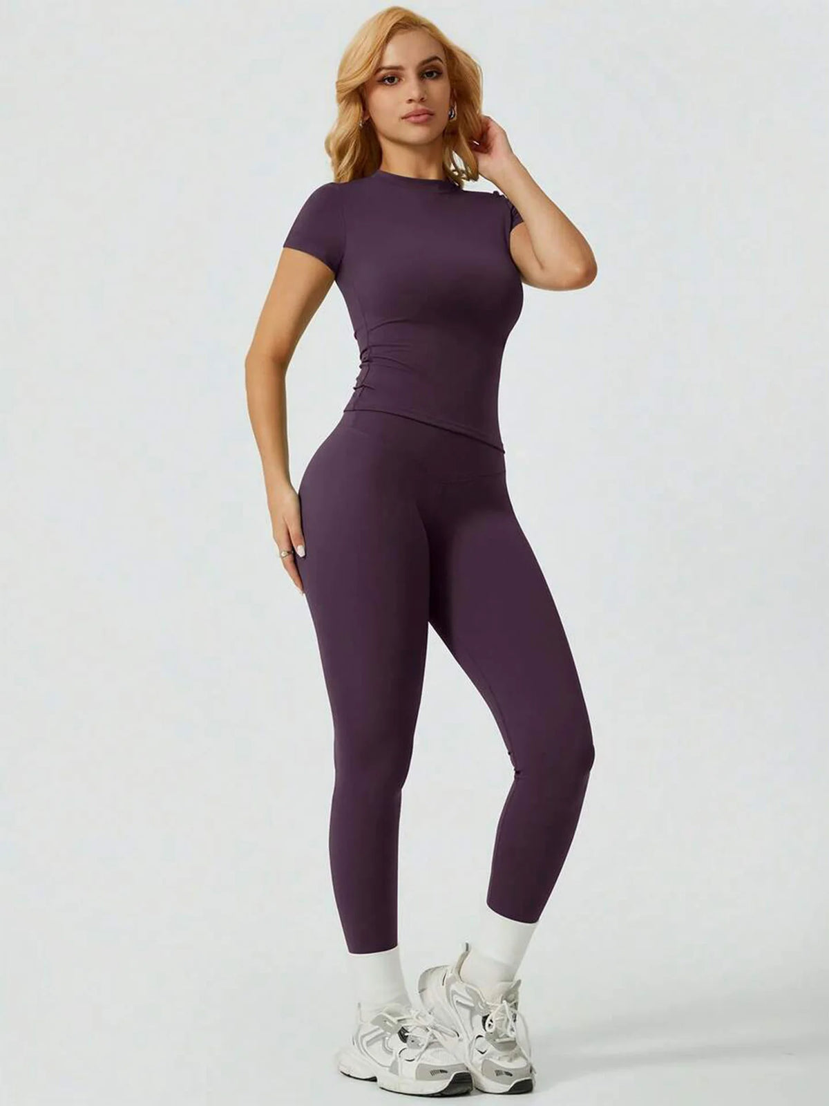 Eine Frau modelliert ein zweiteiliges Yoga-Set mit lila, hoch taillierter Leggings und einem kurzen Oberteil.