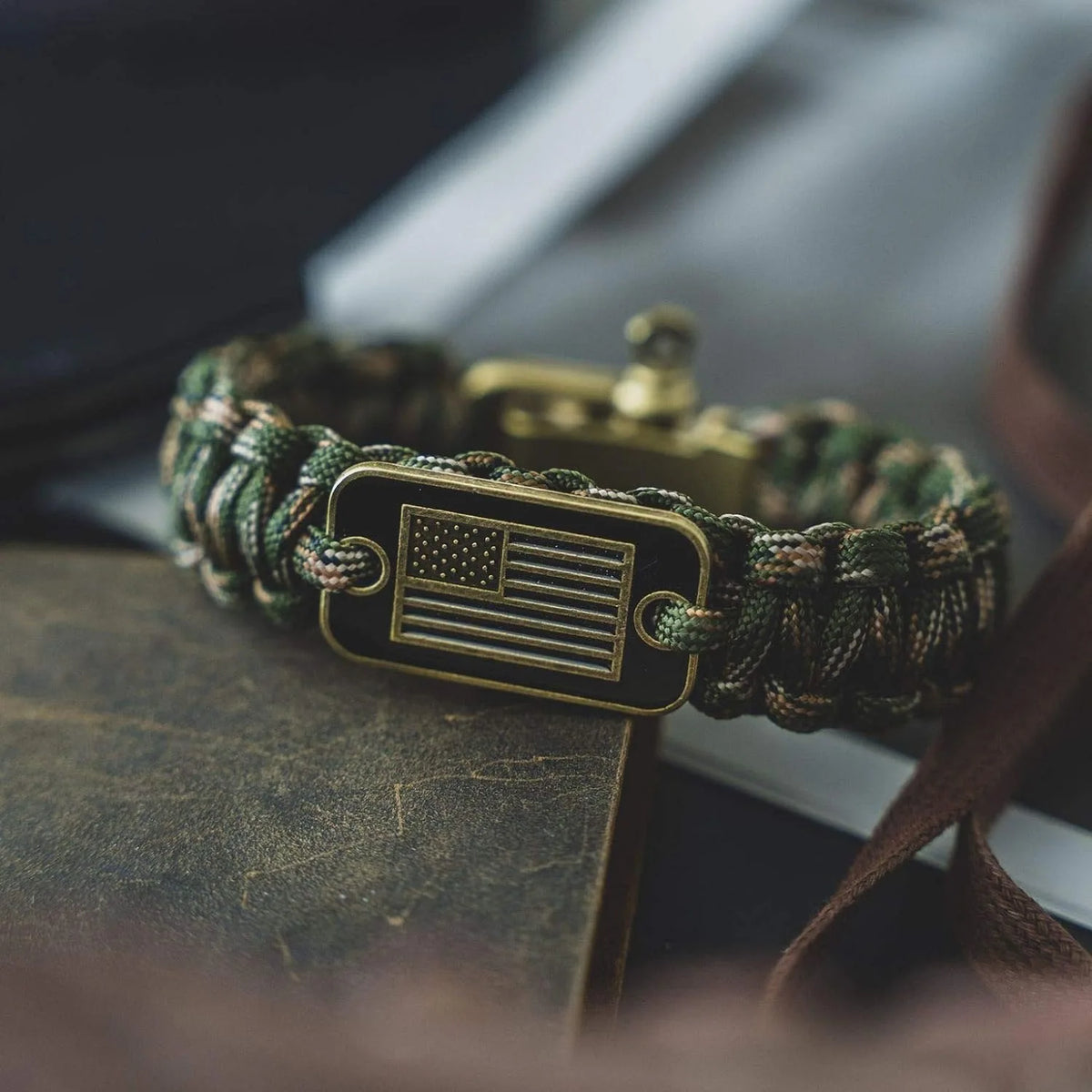 A tactical paracord survival bracelet with a bronze American flag charm resting on a dark surface.