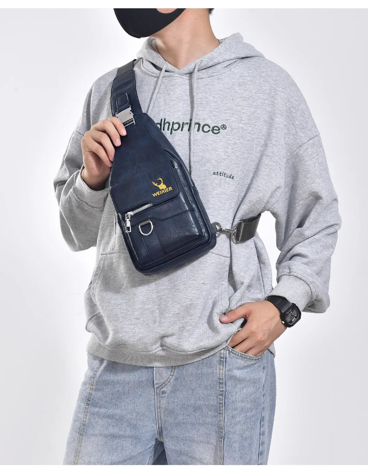 A man wearing a gray hoodie holds a dark blue leather crossbody bag with a gold buckle.