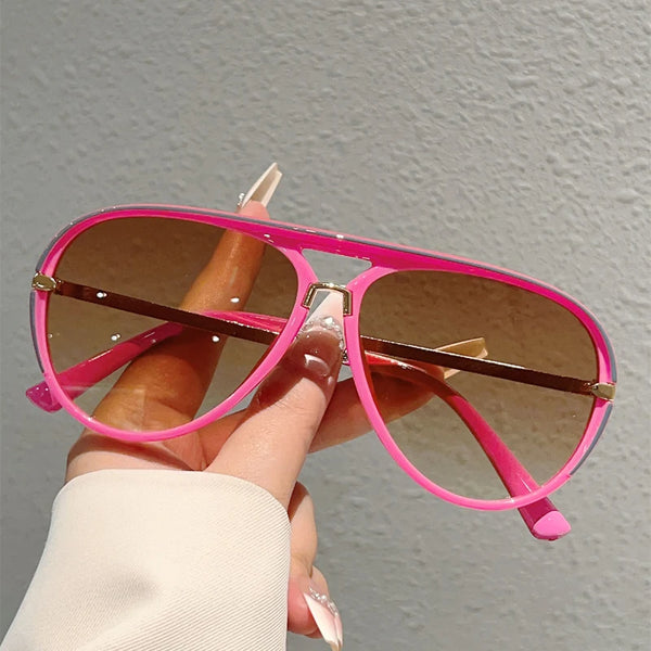 A hand holds a pair of pink vintage pilot sunglasses with gradient lenses against a gray background.