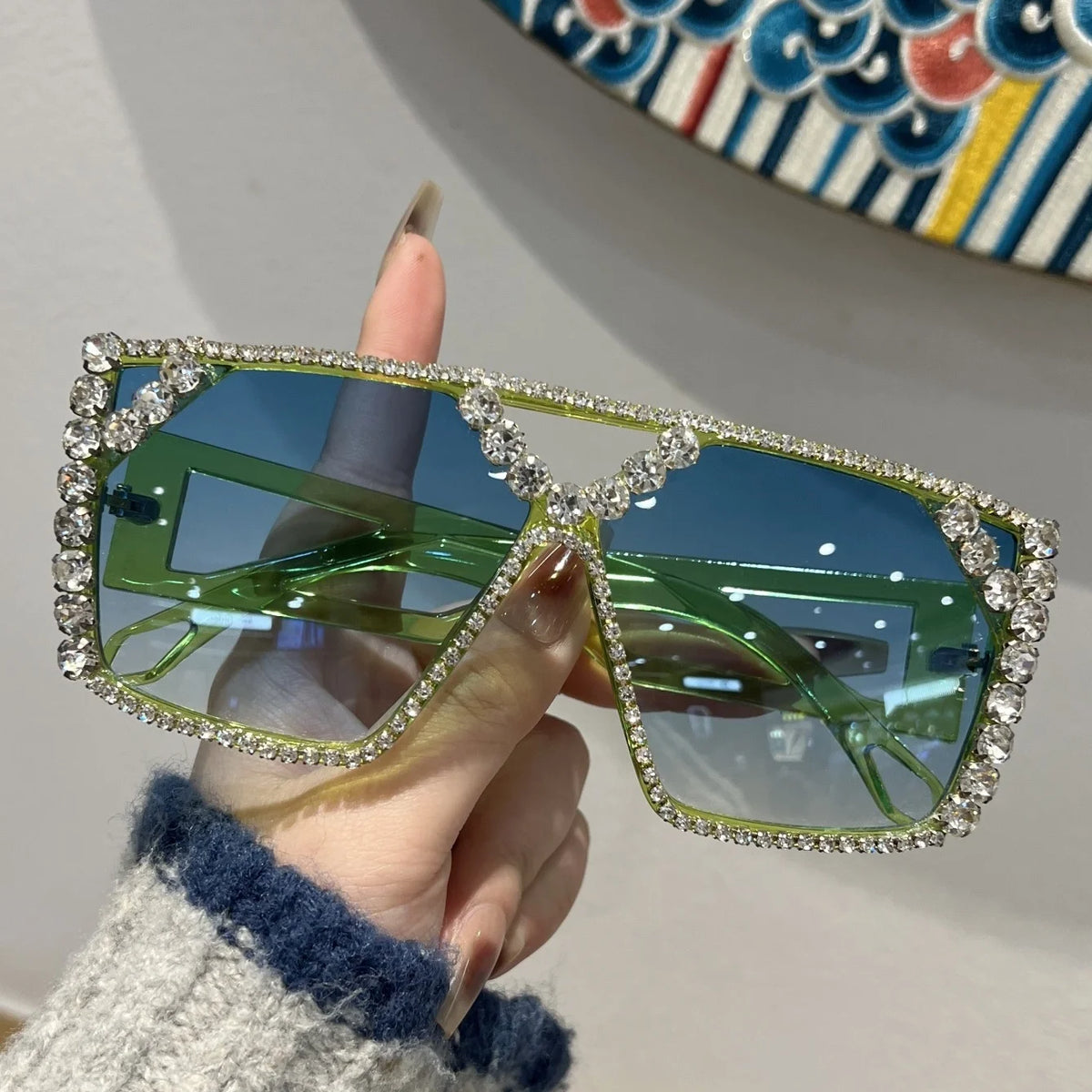 A hand holds a pair of green geometric rhinestone fashion sunglasses against a white background.