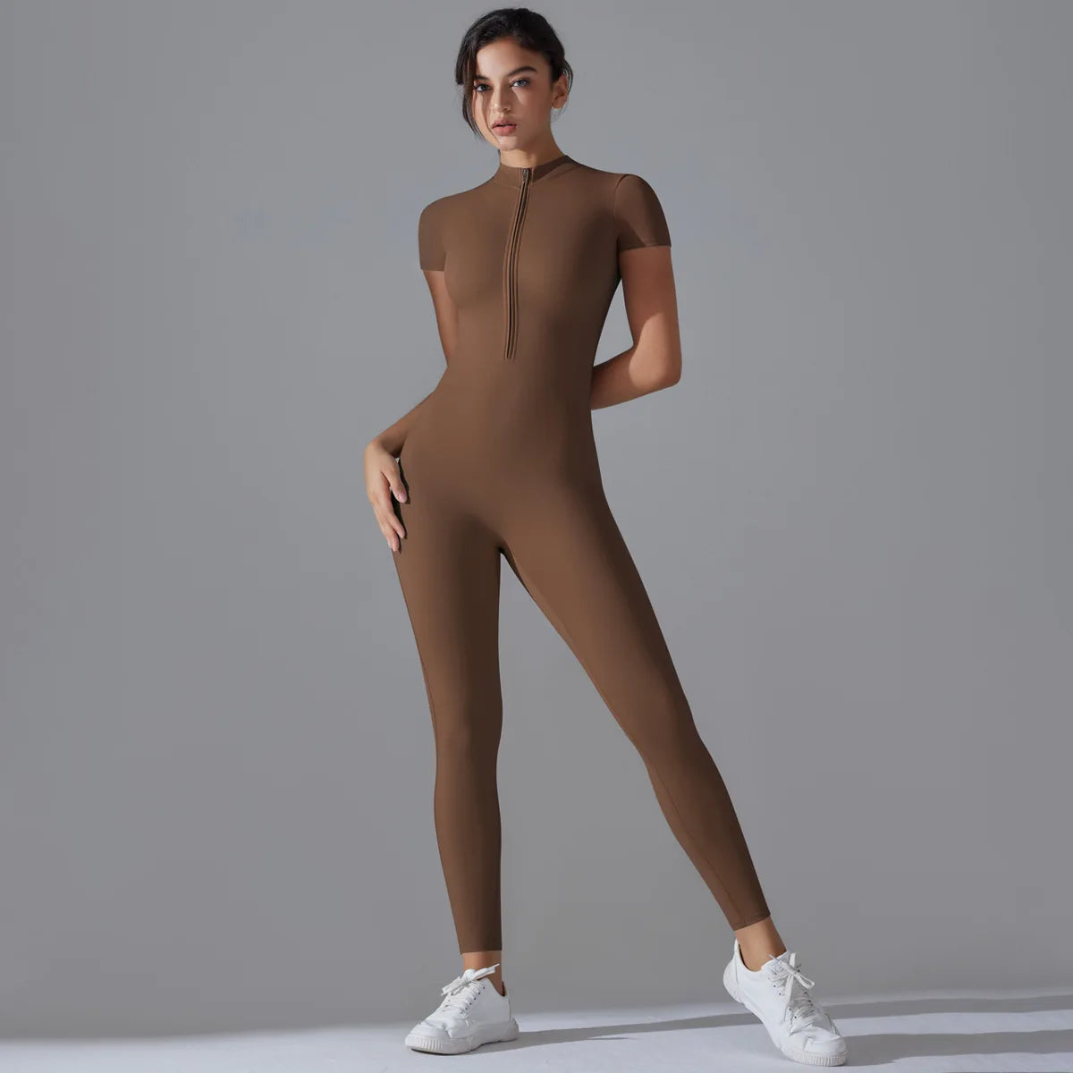 A model wearing a full-length, fitted brown yoga jumpsuit with a high neck zipper and white sneakers.