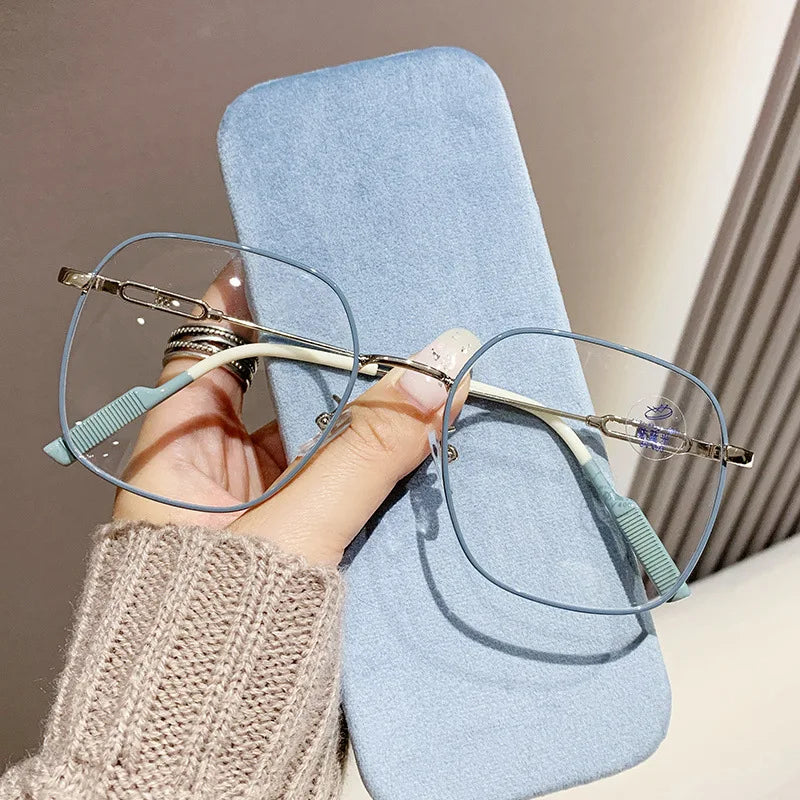 A hand is holding a pair of light blue anti-blue light reading glasses resting on a light blue pad.