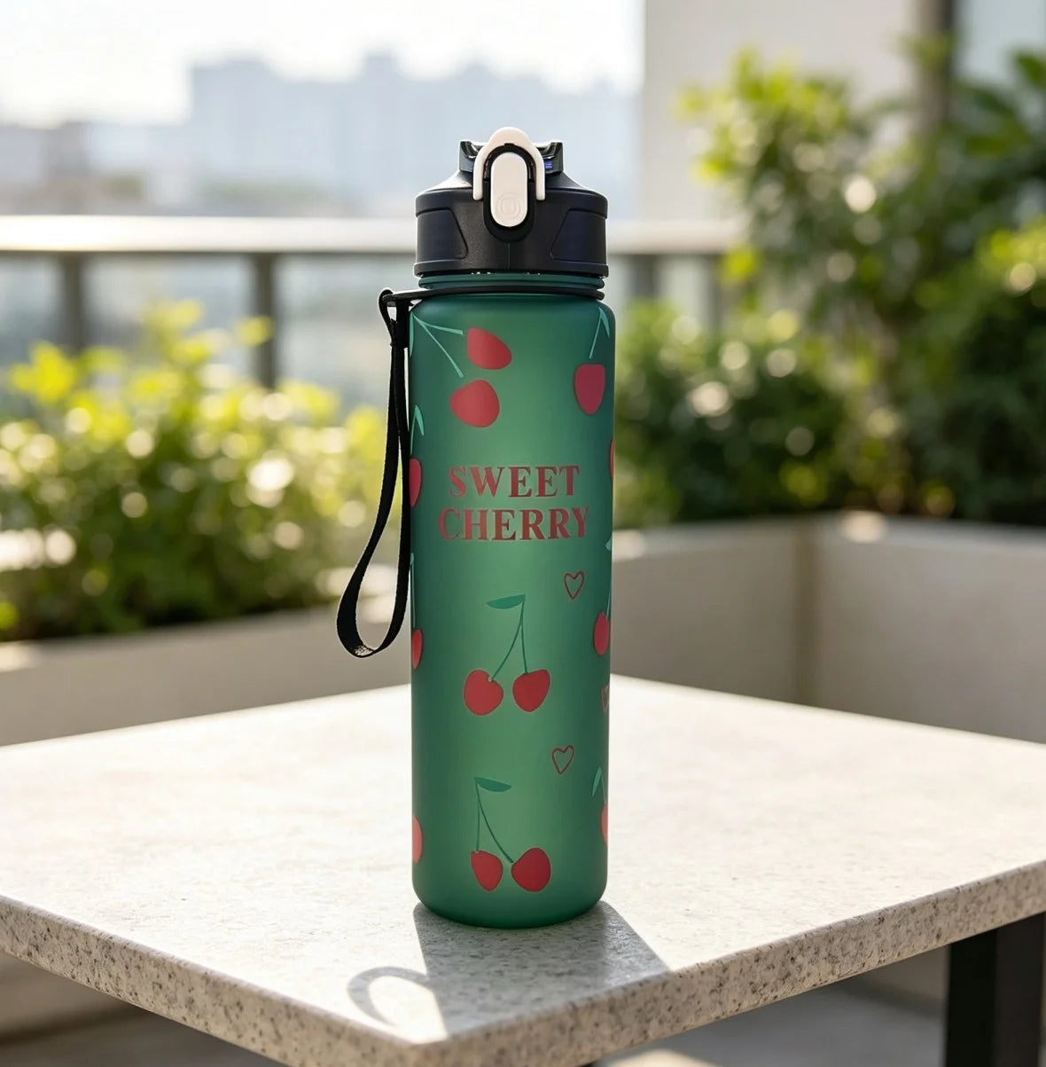 A green reusable water bottle with a sweet cherry design sitting on a light-colored table outdoors.