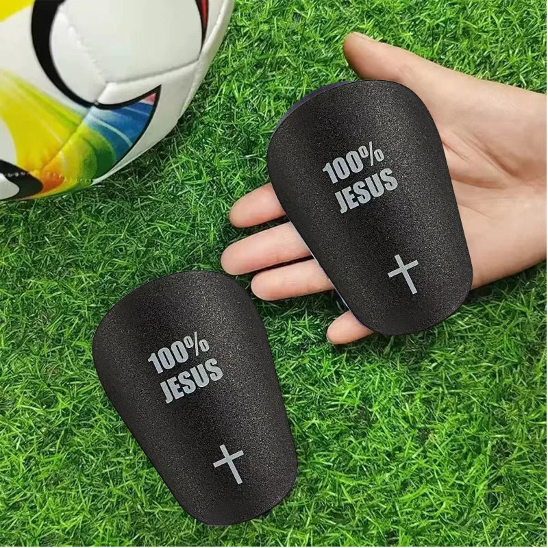 A hand holds a black lightweight soccer shin guard with 100% Jesus printed on it, resting on green grass.