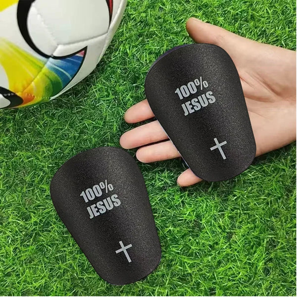 A hand holds a black lightweight soccer shin guard with 100% Jesus printed on it, resting on green grass.