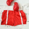 Un par de guantes de boxeo rojos sujetos a una correa de tela blanca, descansando sobre un short deportivo rojo y blanco.
