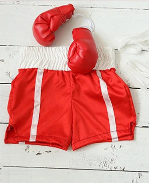 Un par de guantes de boxeo rojos sujetos a una correa de tela blanca, descansando sobre un short deportivo rojo y blanco.