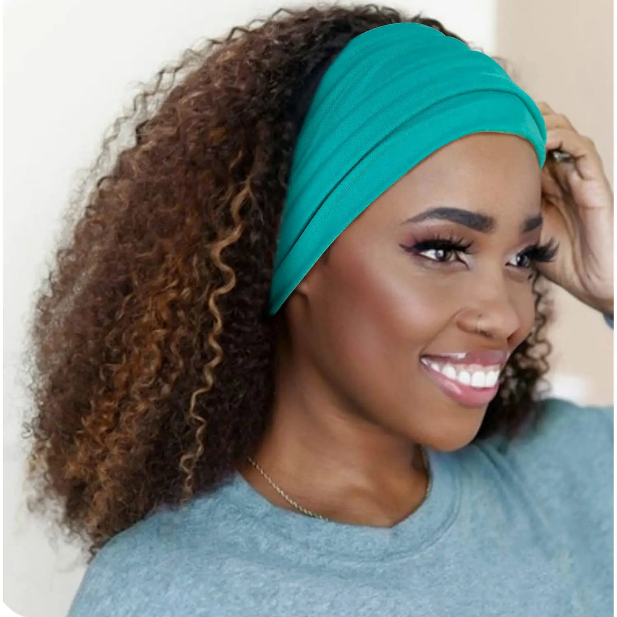 A woman with curly hair is wearing a teal boho wide turban headband and a light blue shirt.