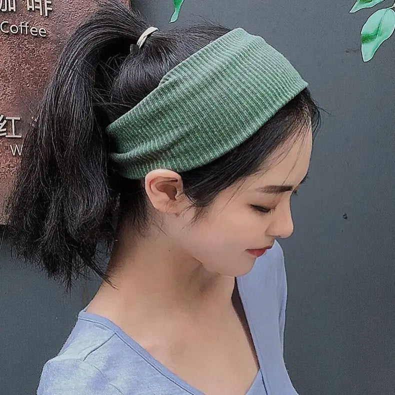 A close-up of a woman wearing a olive green knitted headband on her dark hair, against a dark background.