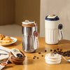 A silver insulated thermos bottle with a white lid and a black tag sits on a wooden table next to a smart display device.