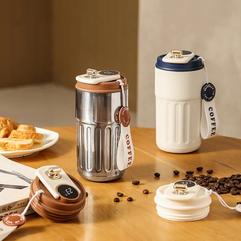 A silver insulated thermos bottle with a white lid and a black tag sits on a wooden table next to a smart display device.