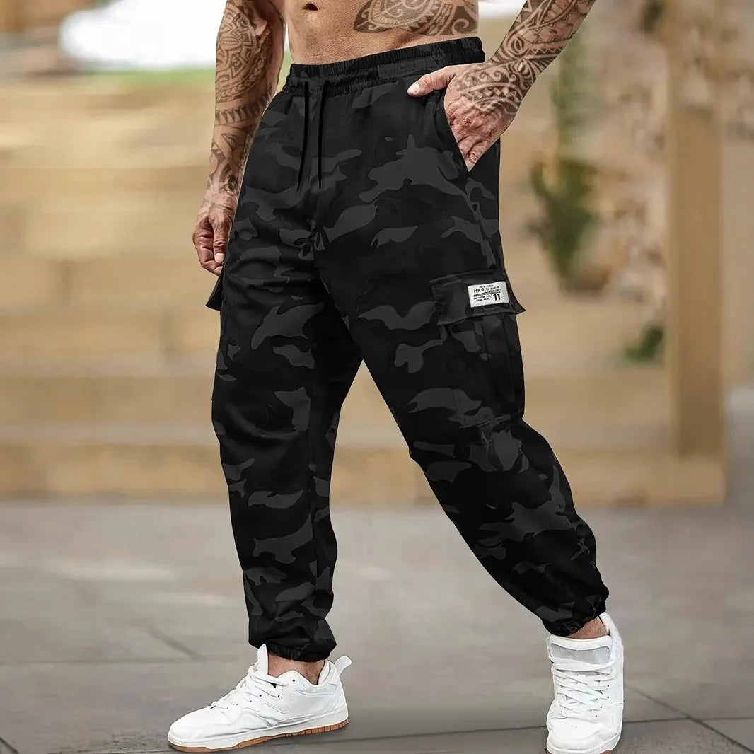 Un homme portant un pantalon cargo camouflage noir et un débardeur, debout à l'extérieur.