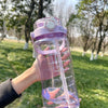 A hand holds a clear purple water bottle with a pink lid and a white straw against a green outdoor background.