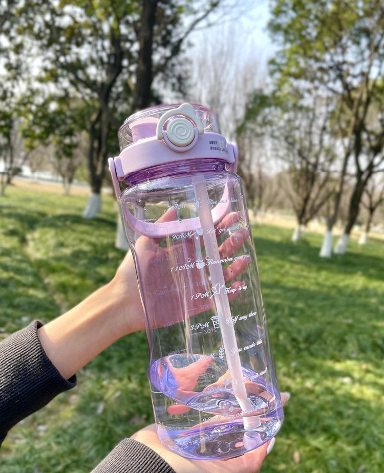 A hand holds a clear purple water bottle with a pink lid and a white straw against a green outdoor background.