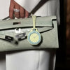 A hand wearing a ring holds a light green leather handbag with a yellow watch charm.