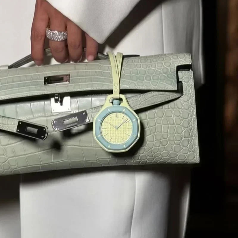 A hand wearing a ring holds a light green leather handbag with a yellow watch charm.