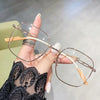A hand holds a pair of thin, wire-framed reading glasses with light-colored frames and orange temples against a blurred backg