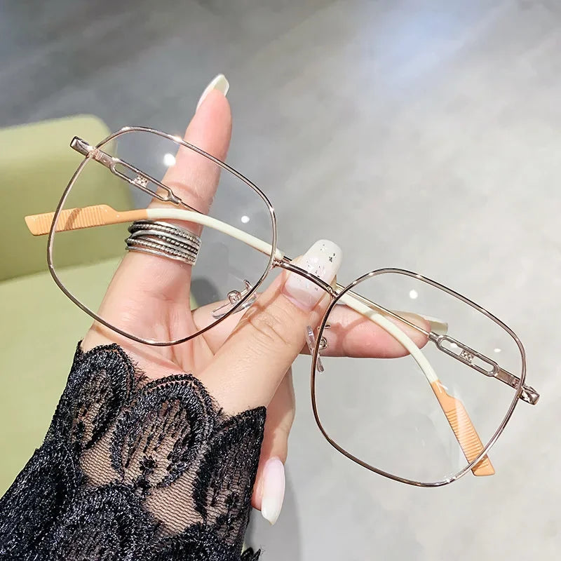 A hand holds a pair of thin, wire-framed reading glasses with light-colored frames and orange temples against a blurred backg