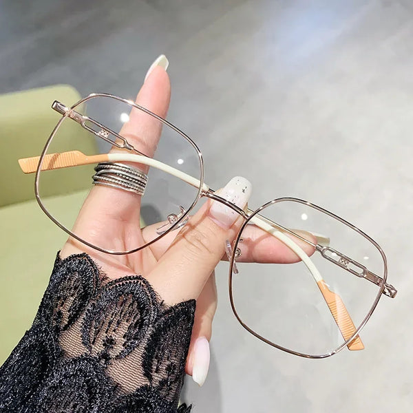 A hand holds a pair of thin, wire-framed reading glasses with light-colored frames and orange temples against a blurred backg