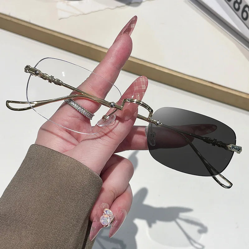 A hand holds a pair of thin, gold-framed rectangle sunglasses with dark lenses against a light background.