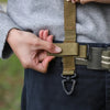 A hand is fastening a tactical nylon glove hook onto a belt loop of dark pants.