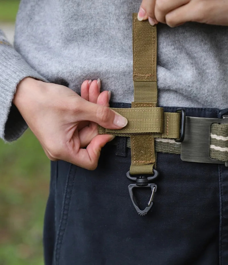 A hand is fastening a tactical nylon glove hook onto a belt loop of dark pants.