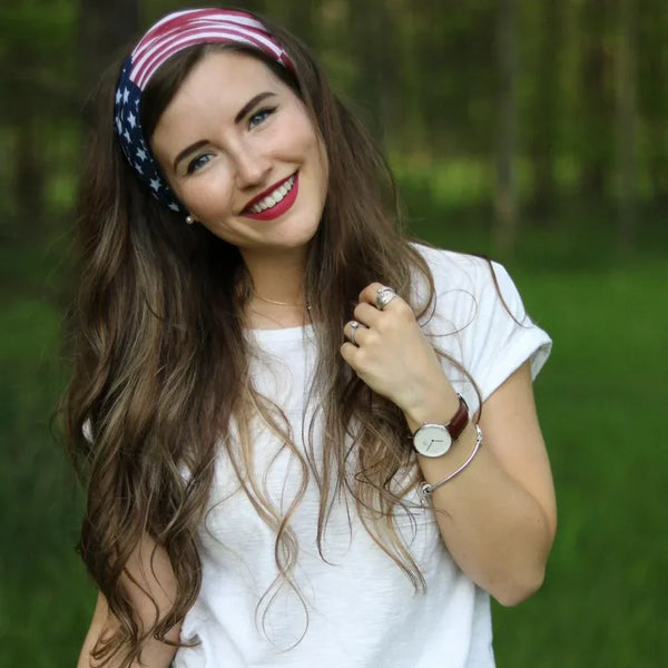 Une jeune femme aux longs cheveux bruns portant un bandana et foulard du drapeau américain, souriante à l'extérieur.