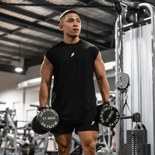 A muscular man wearing a black sleeveless fitness tank and shorts is standing in a gym setting.