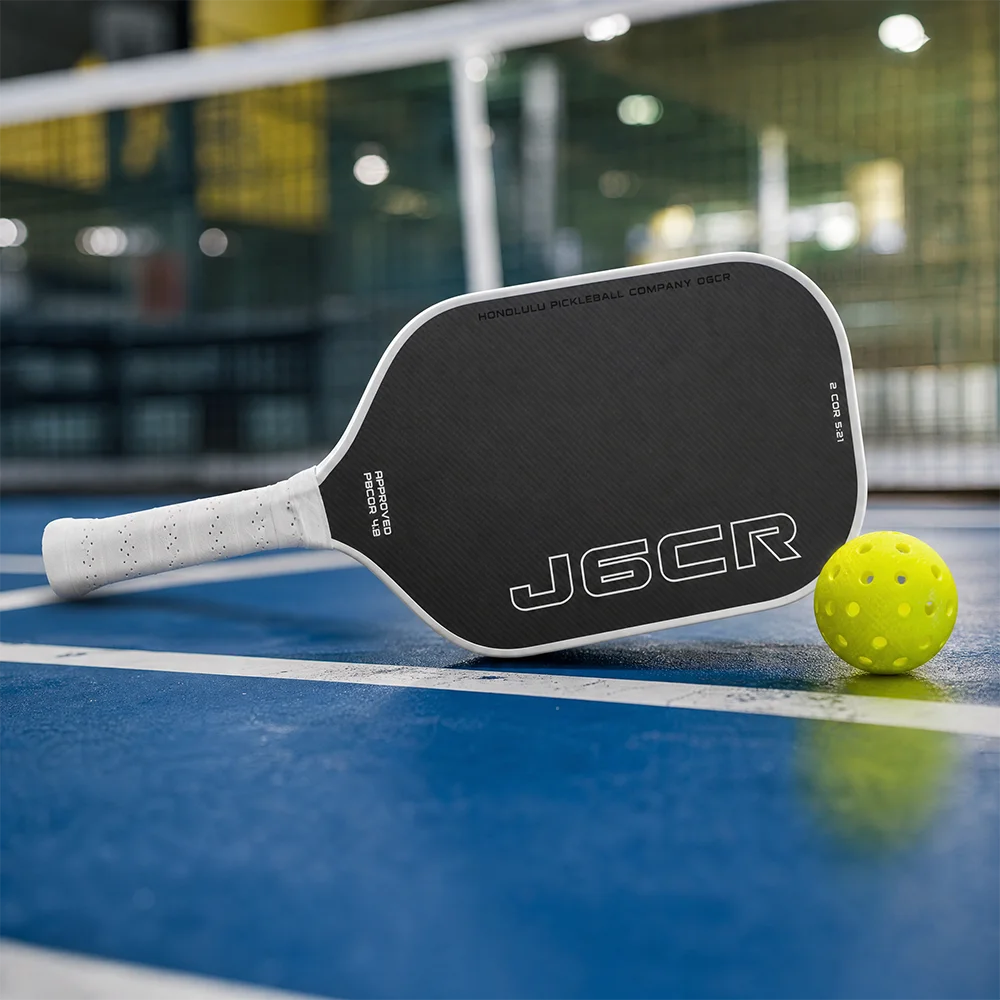 Schwarzes Carbonfaser-Pickleball-Schläger auf blauem Platz mit einem gelben Pickleball.
