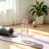 Eine klare Wasserflasche mit rosa Deckel und Zeitmarkierung steht auf einer lila Yogamatte neben einer Topfpflanze.