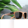A pair of light tan polarized square sunglasses resting on a white surface with green foliage in the background.