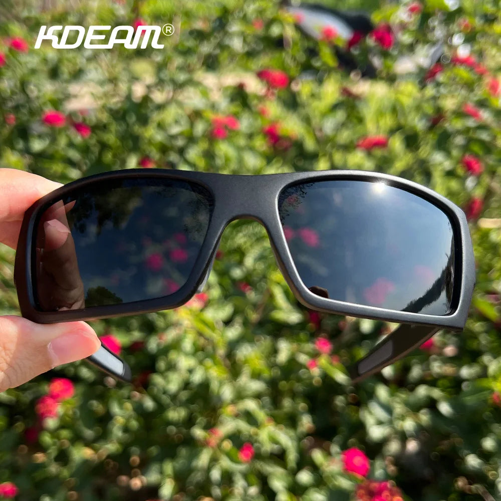 A pair of black polarized sports sunglasses being held against a blurred background of green foliage and red flowers.