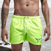 A muscular man wearing neon yellow quick-drying swim shorts standing near the ocean.