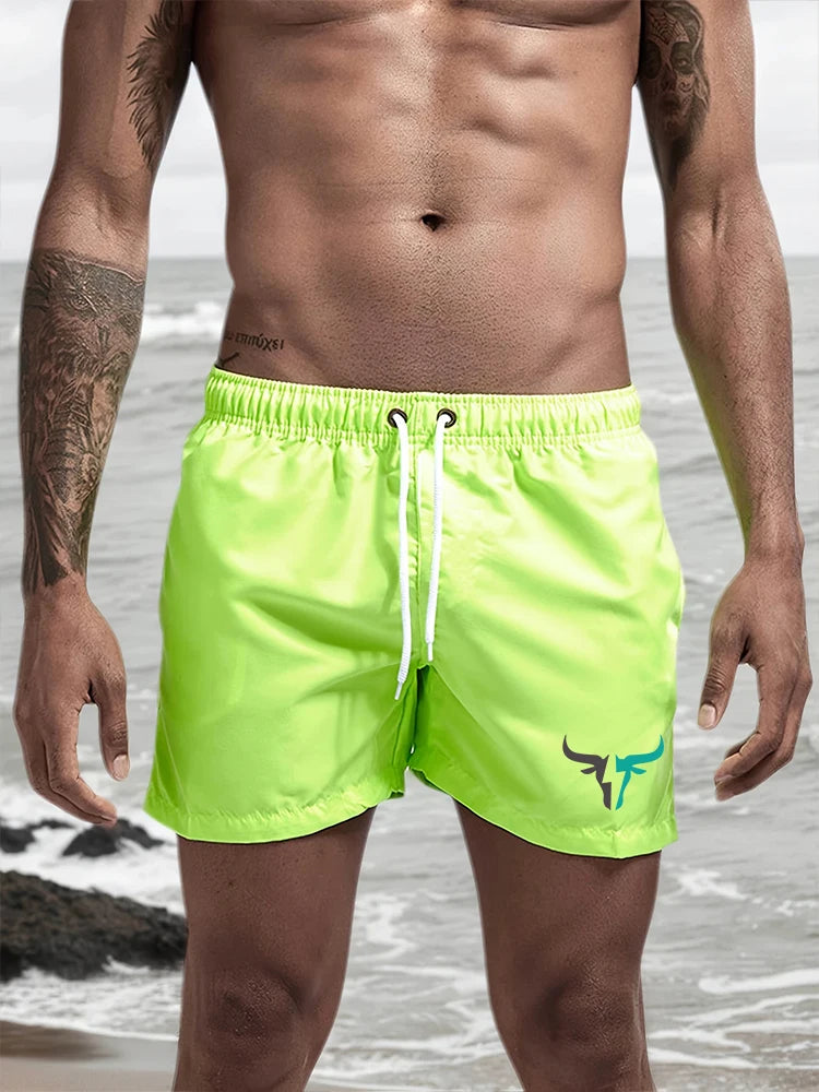 A muscular man wearing neon yellow quick-drying swim shorts standing near the ocean.